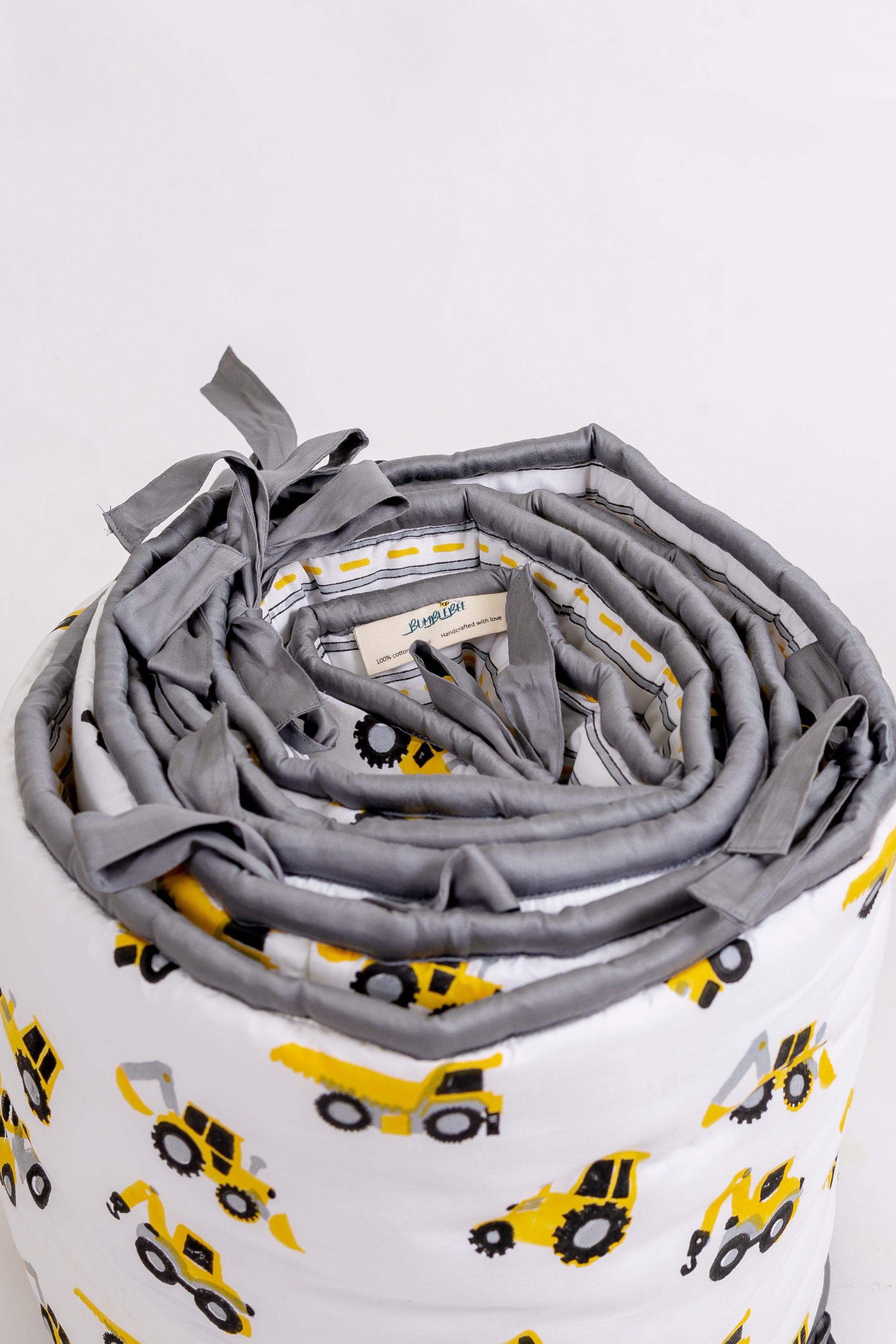 Top view of rolled construction truck crib bumper showing grey fabric ties, striped reverse and Bumblebee label